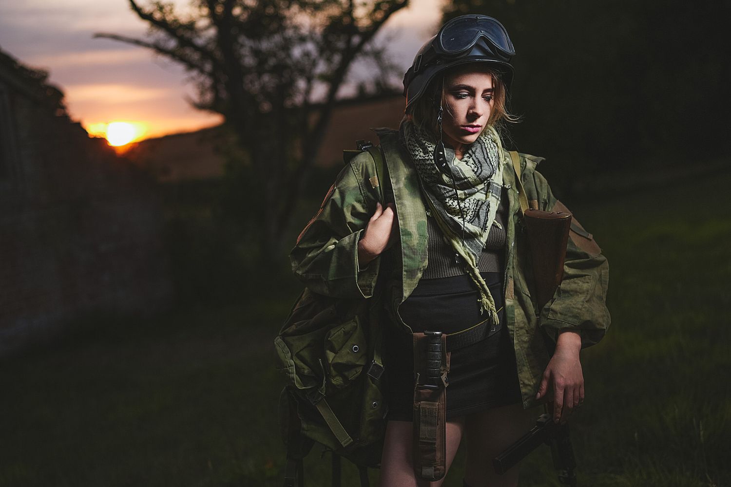 Woman in military gear and helmet poses at sunset wearing olive jacket and scarf in dramatic outdoor setting.