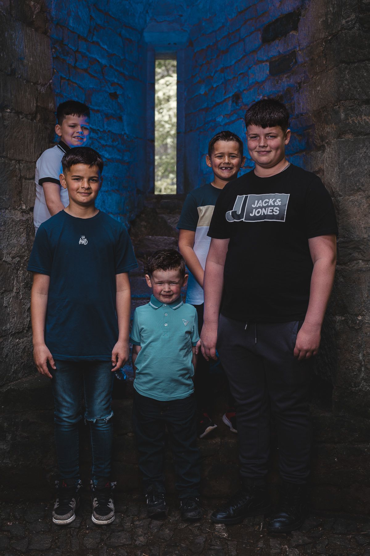 Five boys posing together in a stone archway with blue lighting, wearing casual clothes including a Jack & Jones shirt.