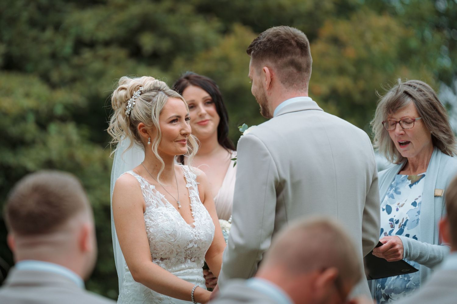 Bride and groom exchange vows outdoors at a wedding ceremony, with smiling guests and officiant nearby.