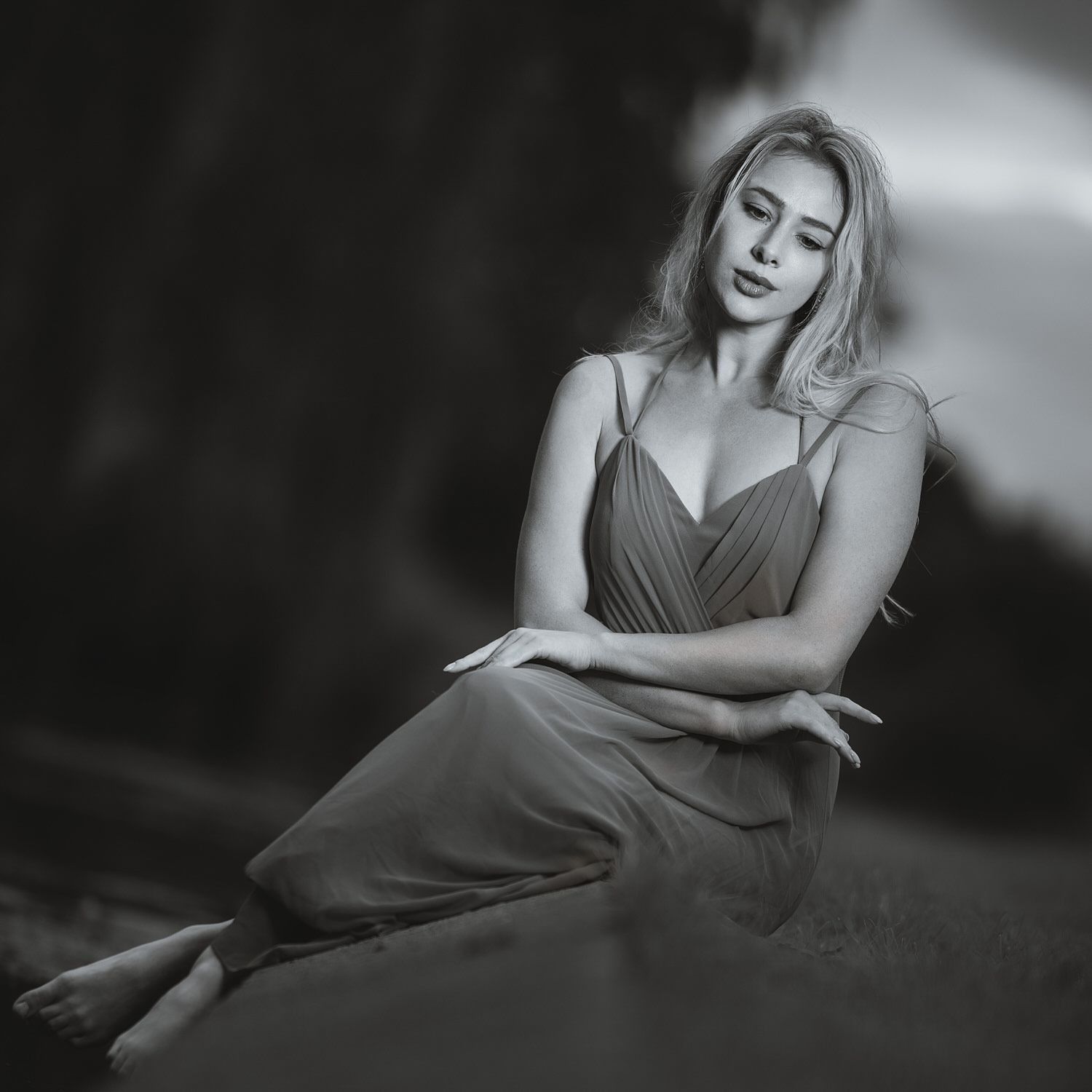 Black and white portrait of a blonde woman in a spaghetti strap dress sitting gracefully outdoors.
