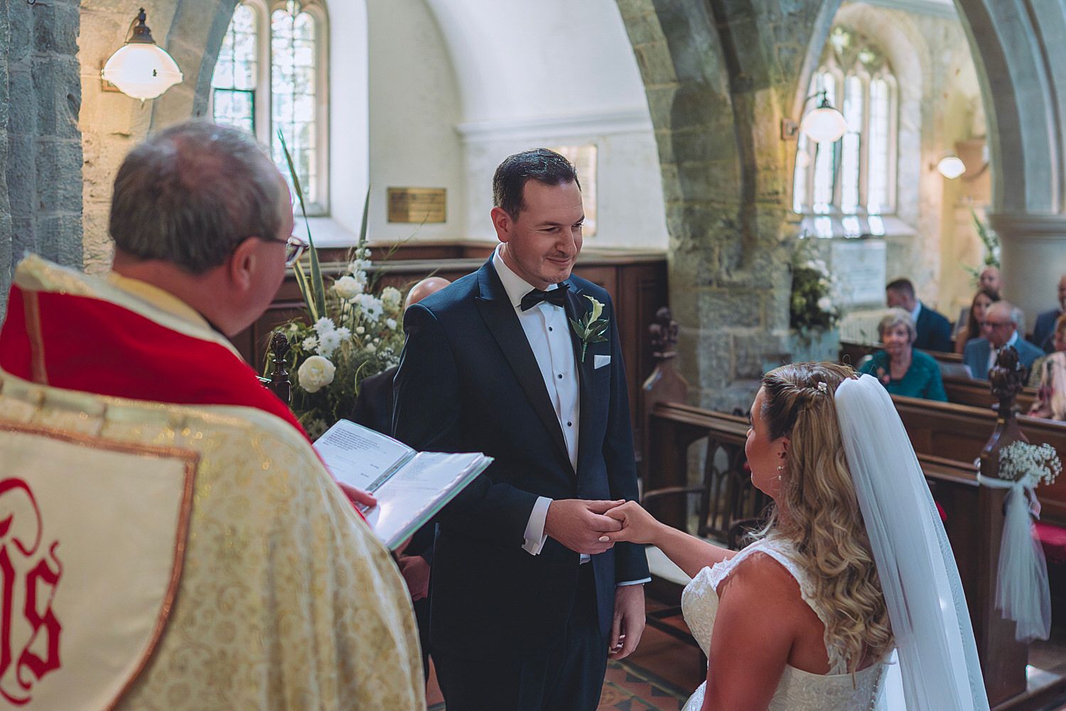 Groom exchanges vows with bride at church altar, officiant in red vestments holding ceremony book.