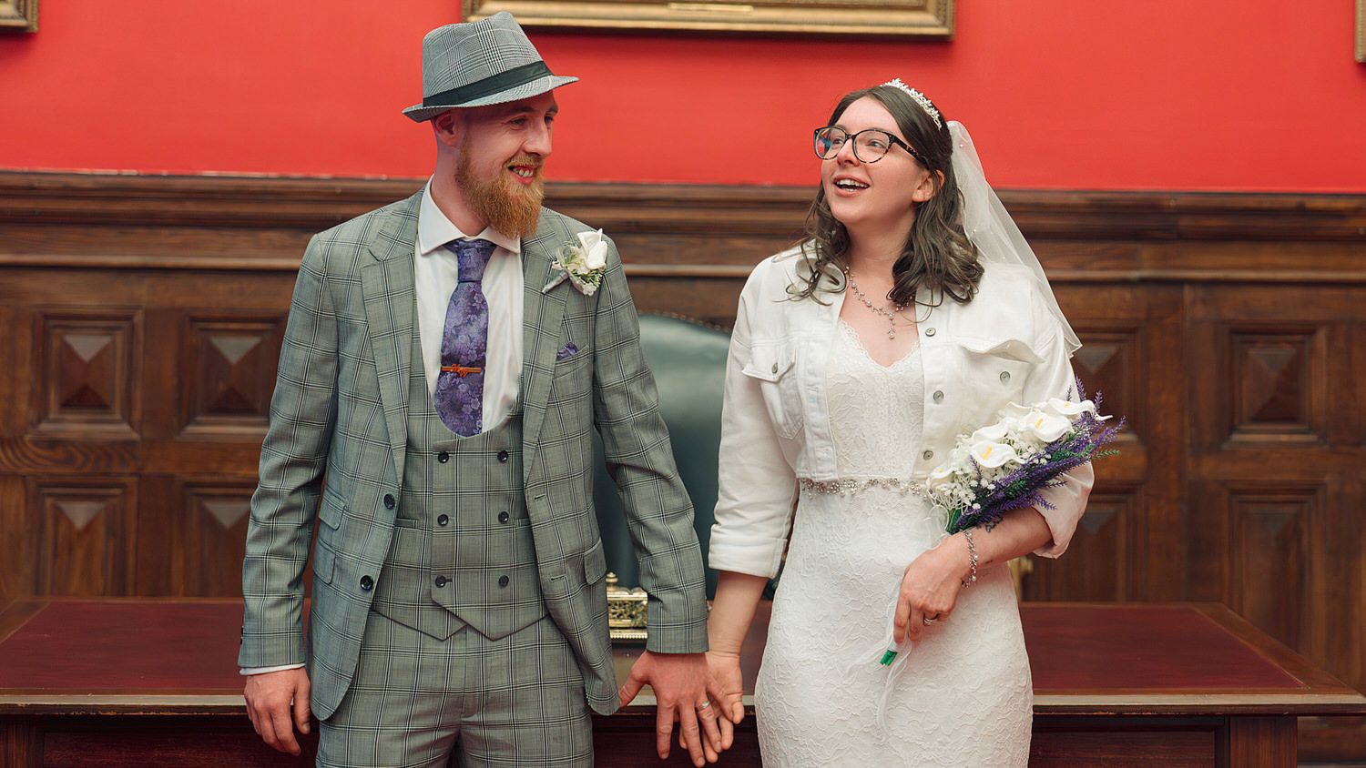 A smiling bride and groom hold hands in a registry office, she in white lace, he in a grey tweed suit and hat.