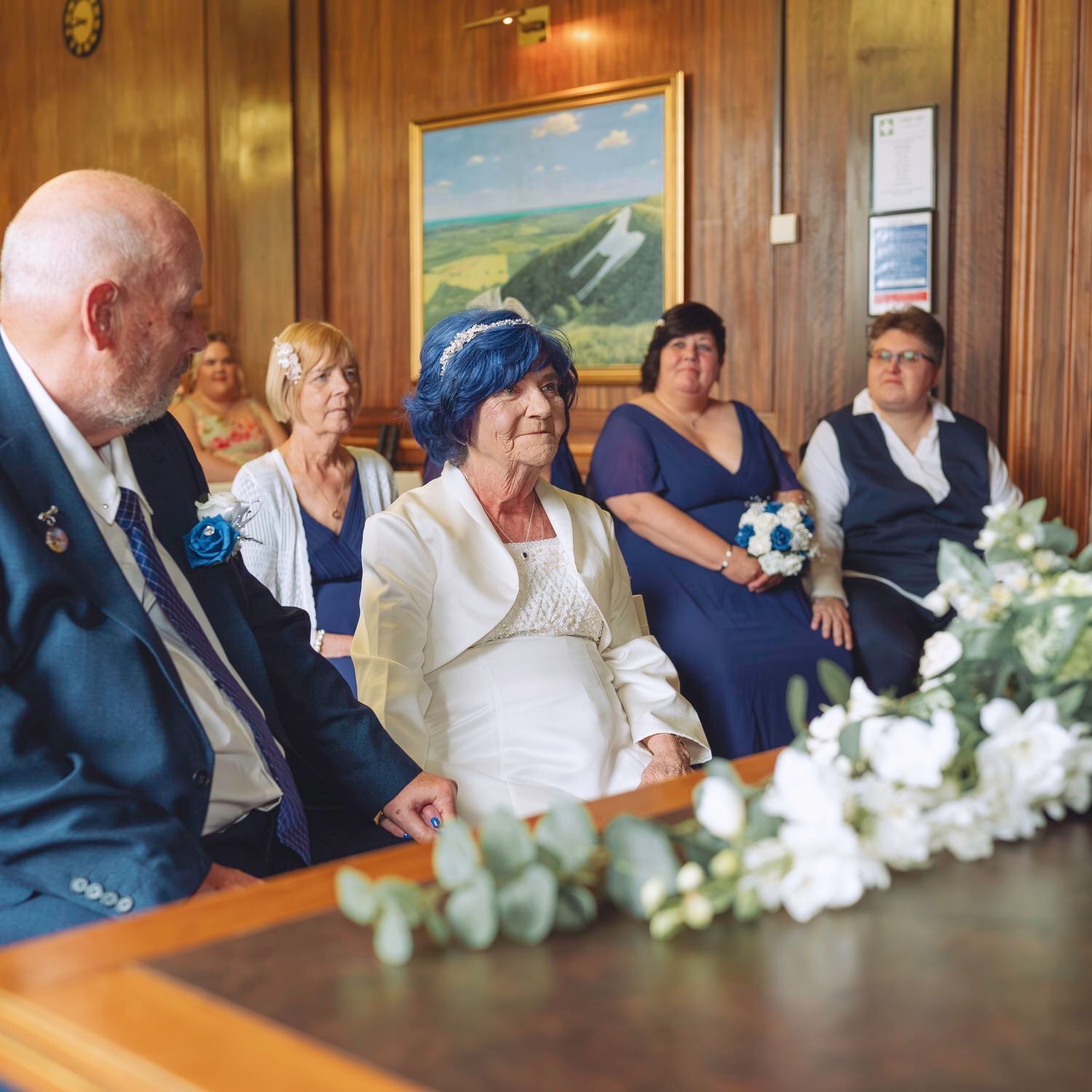 Elderly couple exchanging vows at a registry office wedding ceremony, with bridesmaids in blue dresses seated behind them.