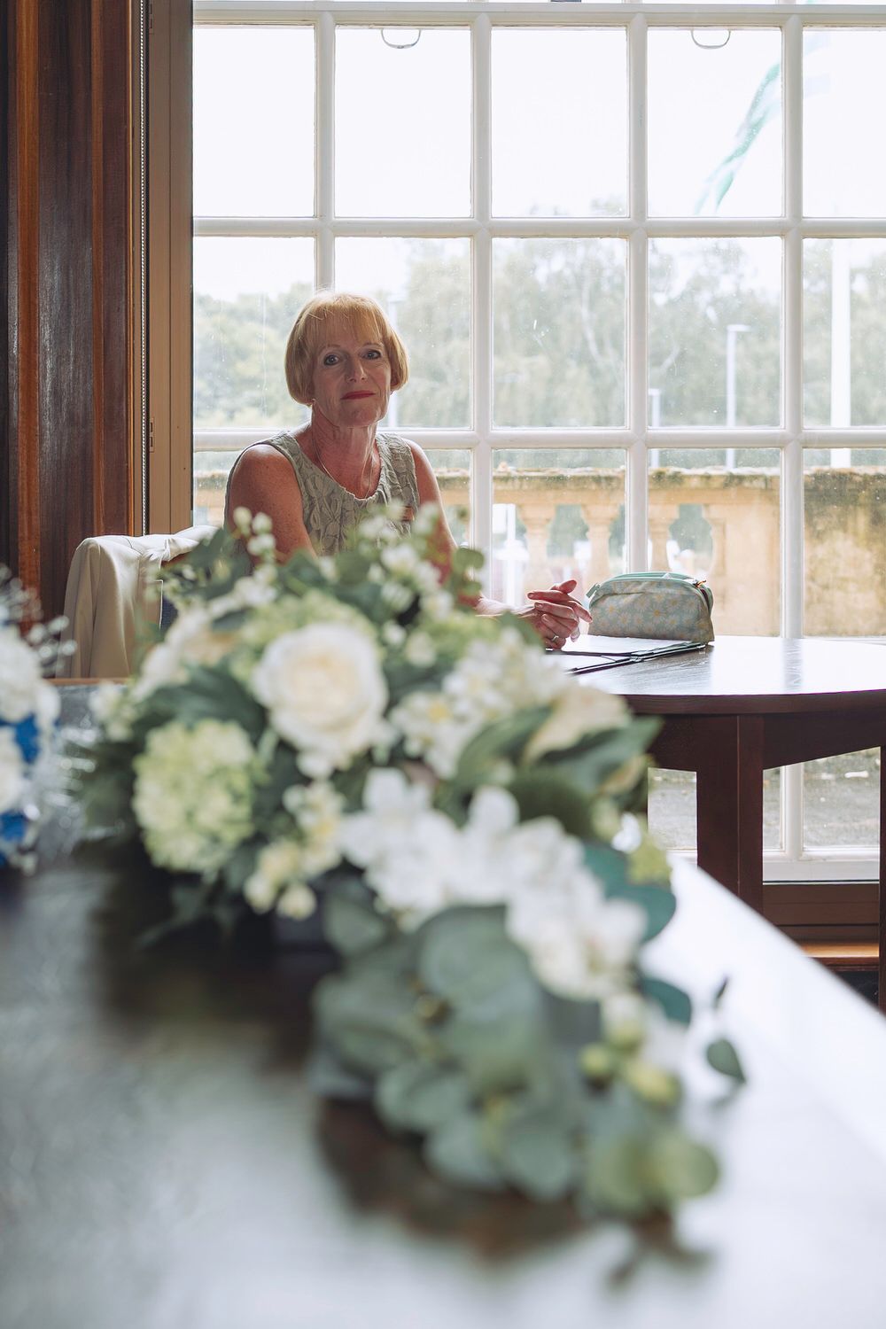 Elegant woman seated behind a floral arrangement of white roses and greenery at a wedding reception table.
