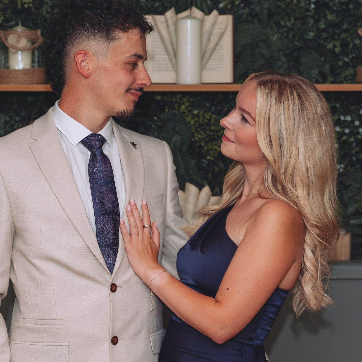 Couple in formal attire gazing at each other, man in beige suit with navy tie, woman in navy dress with blonde hair.