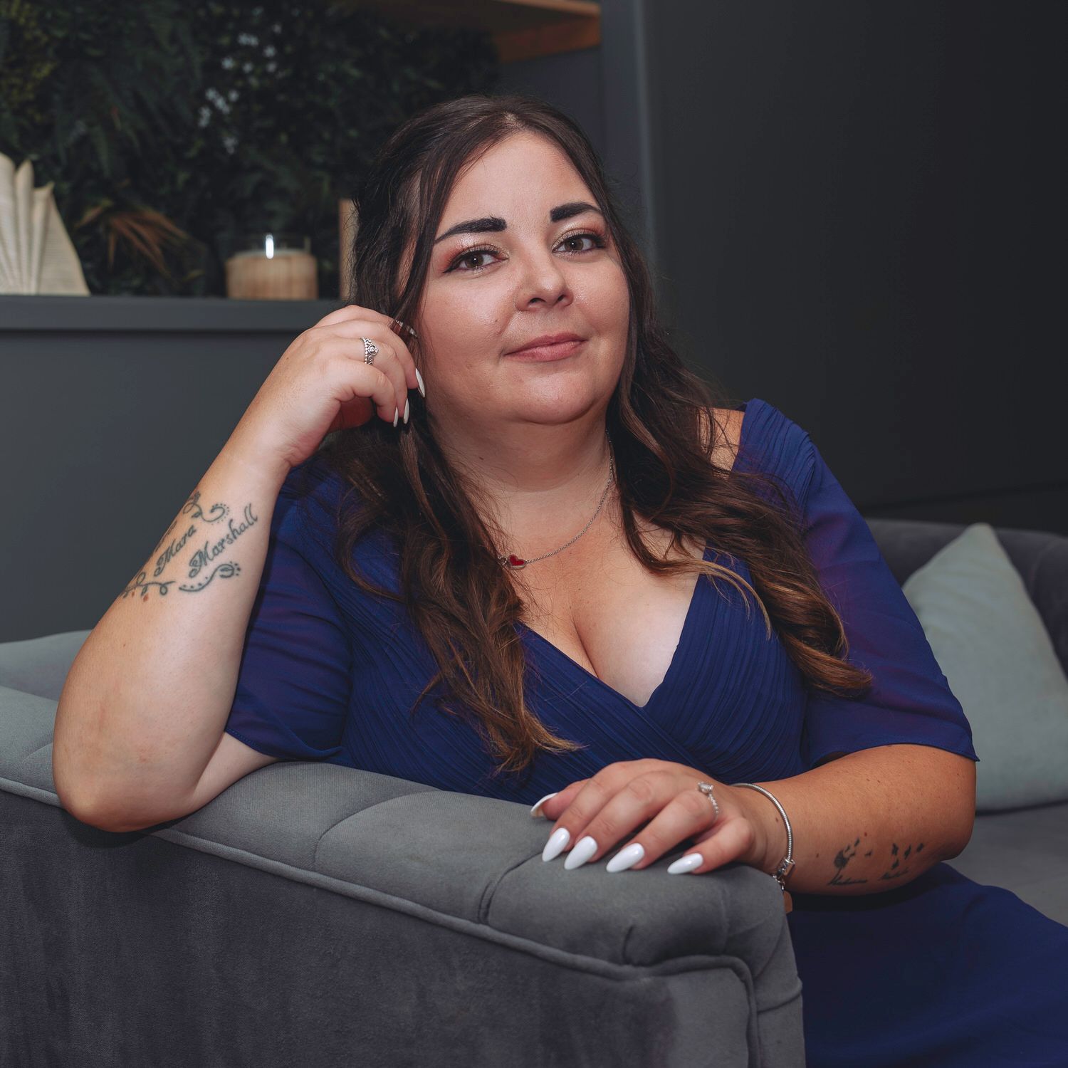 A woman with long brown hair and tattoos sits on a gray sofa wearing a blue dress with white manicured nails.