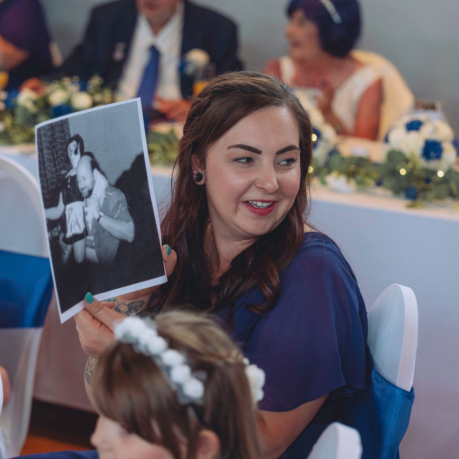 Woman in blue dress holding a black and white photo at a wedding reception with floral decorations.