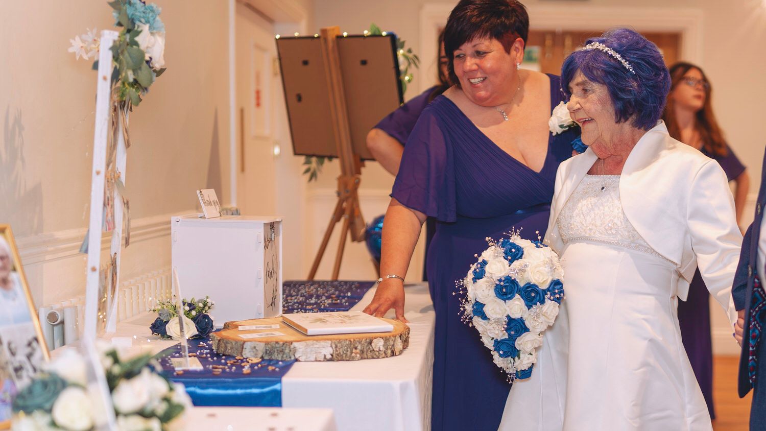 Two women in formal attire smile at a wedding table adorned with blue and white floral decorations.