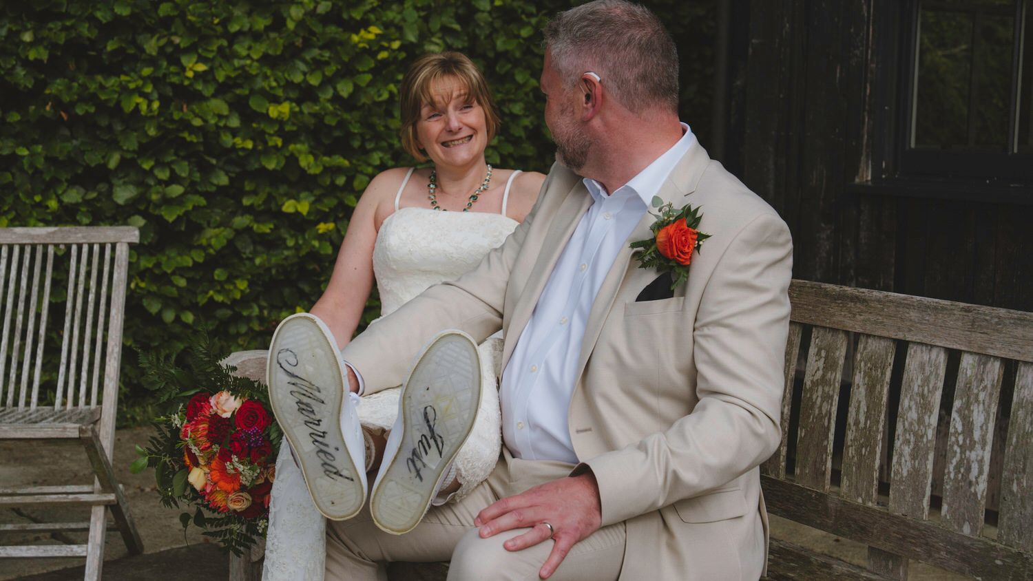 Bride and groom sitting on a wooden bench, smiling, bride showing embroidered wedding shoes, groom in tan suit with boutonniere.