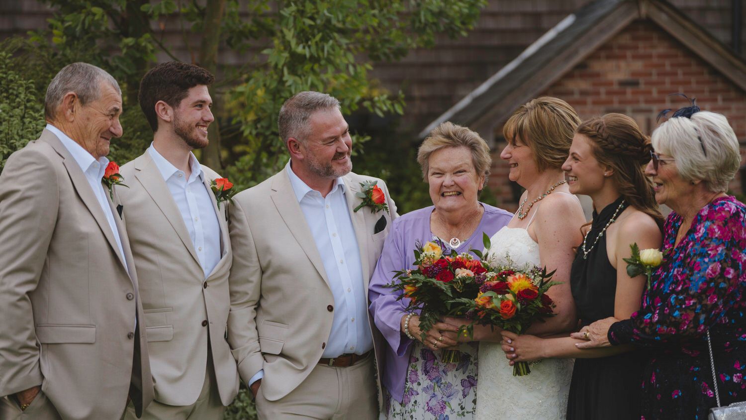 Wedding party smiling outdoors, bride holding colorful bouquet, surrounded by family in elegant attire near brick building.
