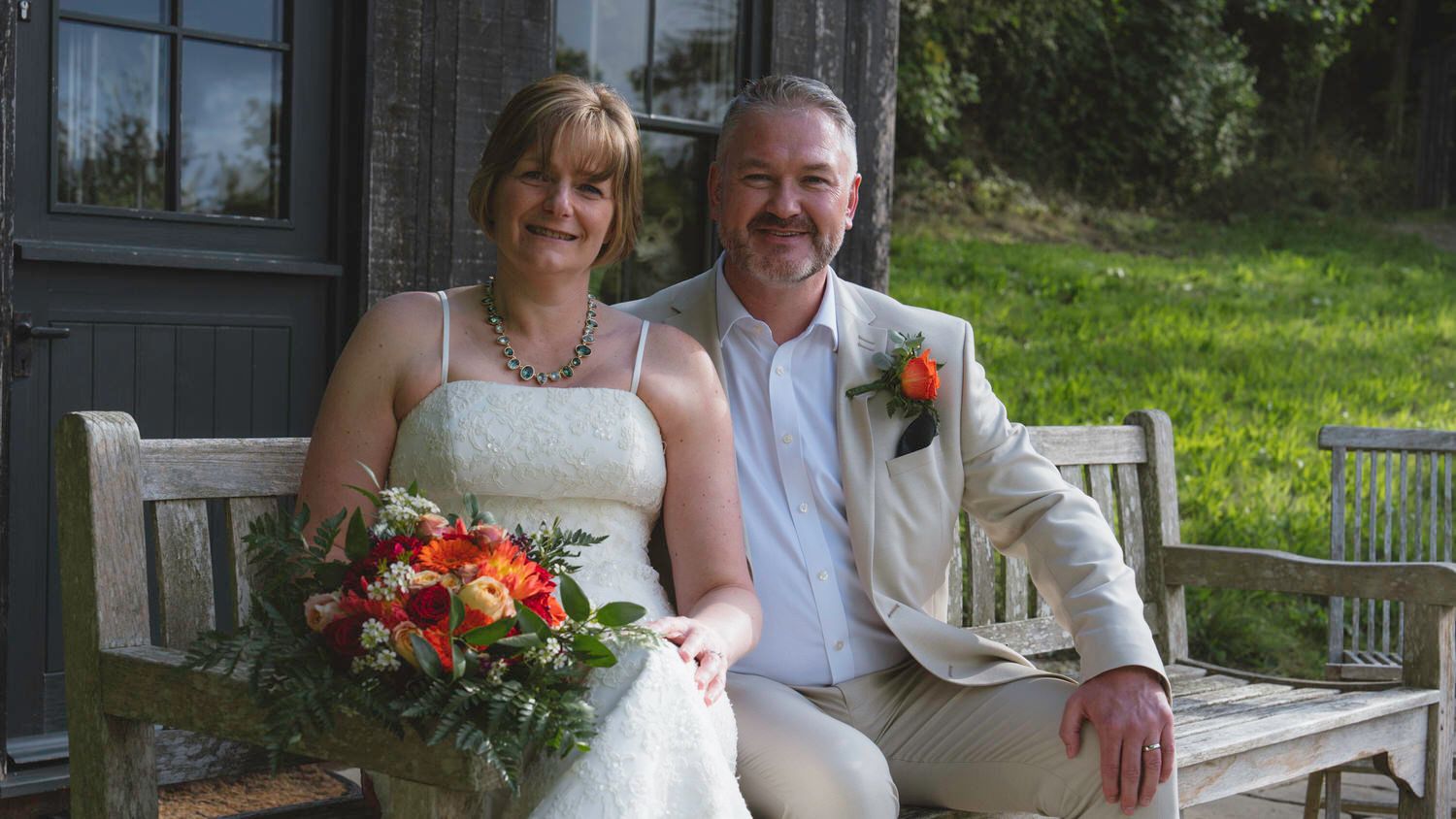 Bride and groom sitting on a wooden bench outdoors, bride holding a colorful bouquet, groom in a beige suit.