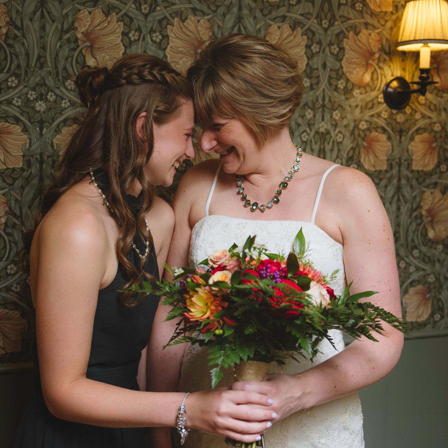 Bride in white dress holding colorful bouquet shares tender moment with bridesmaid in black dress at wedding.