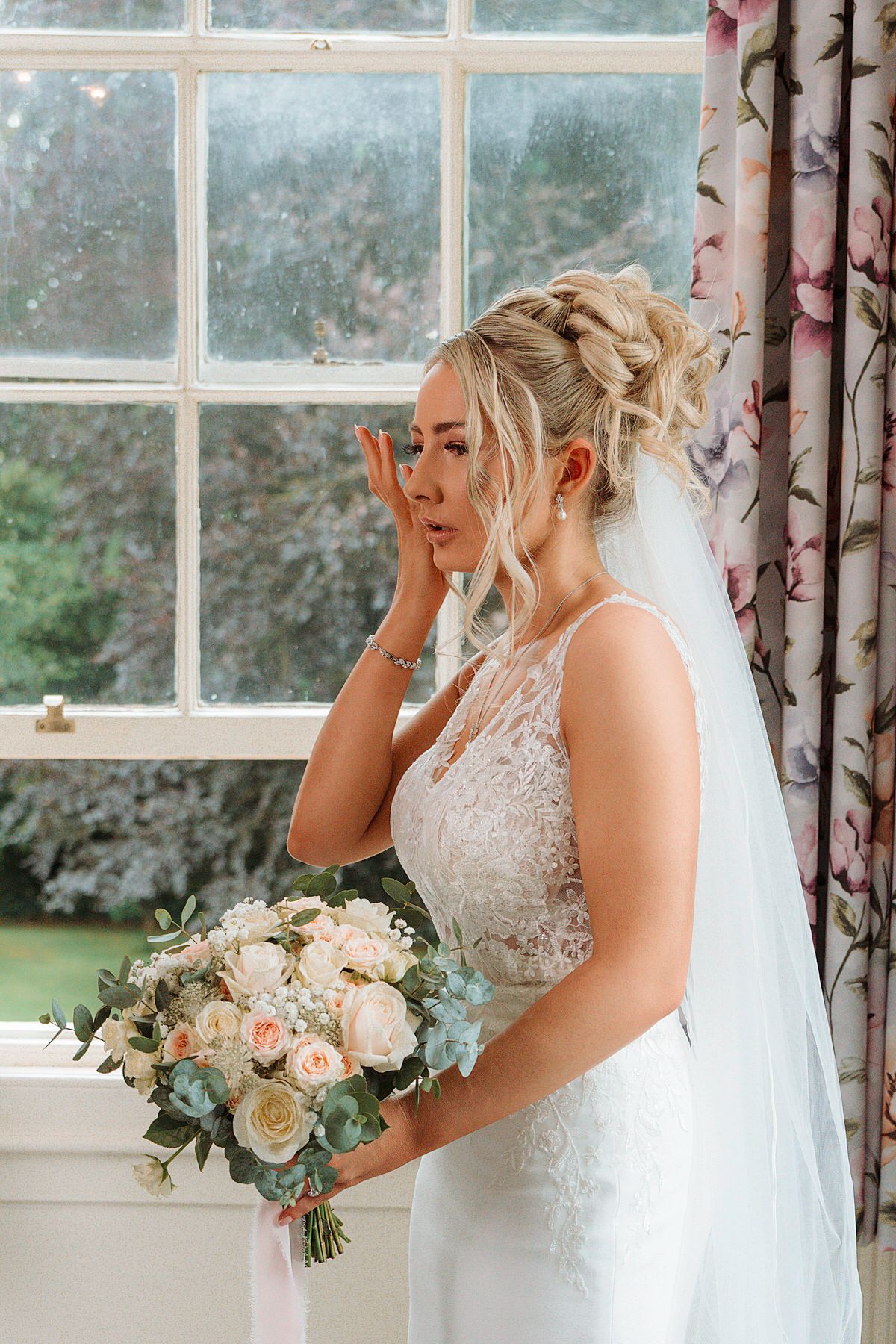 Emotional bride in lace wedding gown holding bouquet of roses near window, wiping away tears.