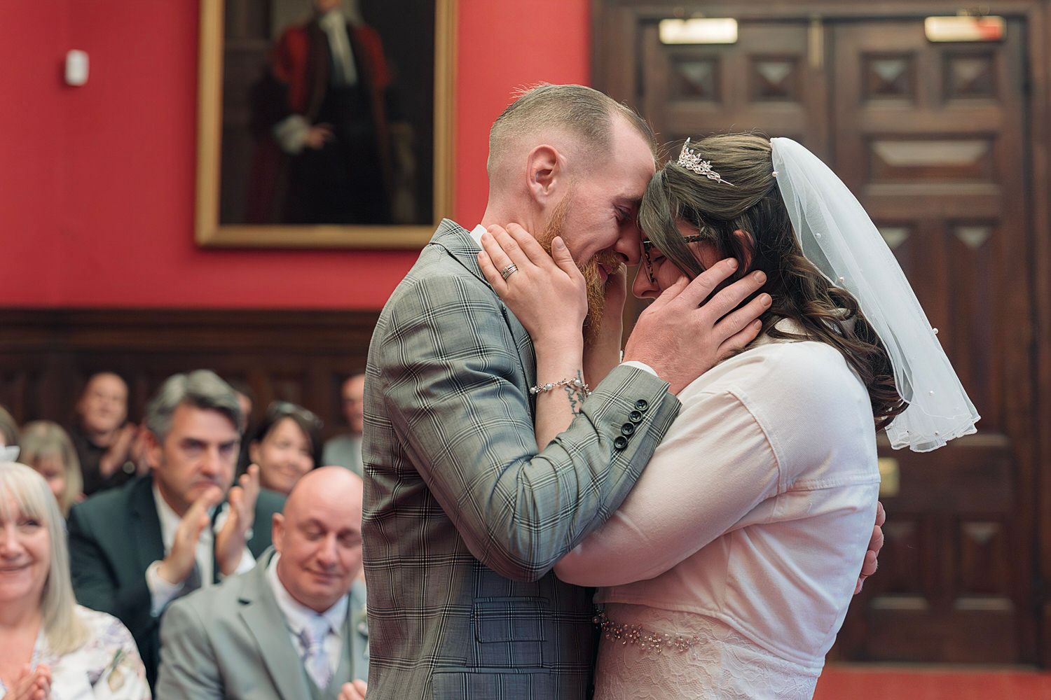 Bride and groom share their first kiss at a wedding ceremony in an elegant red-walled venue with guests applauding.