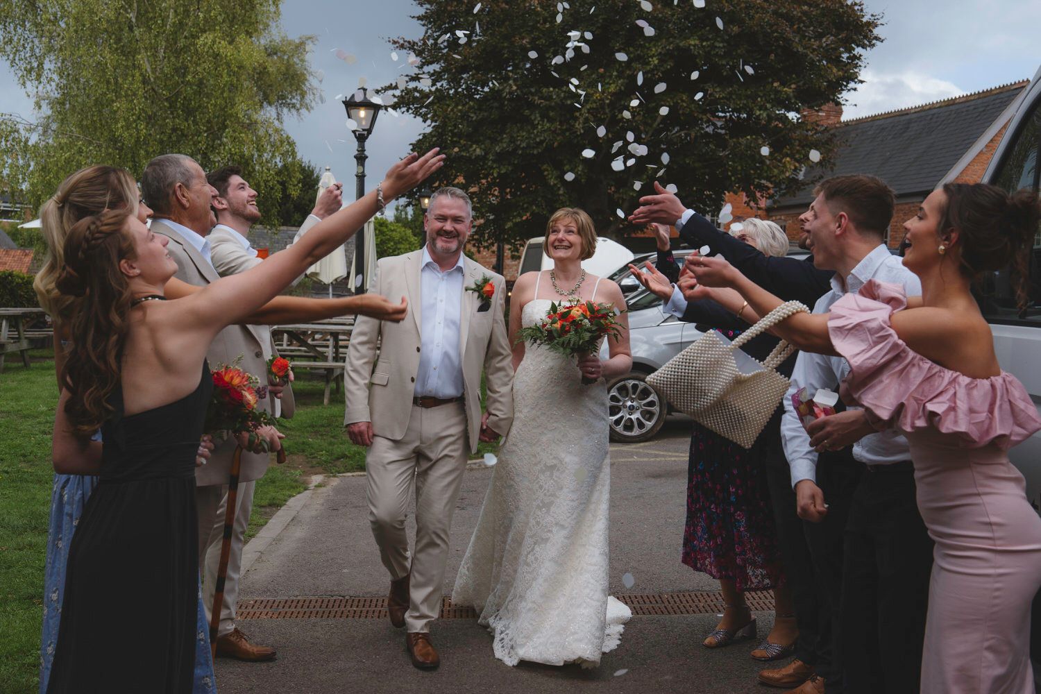 Smiling newlyweds walk through confetti tunnel of cheering wedding guests outdoors.