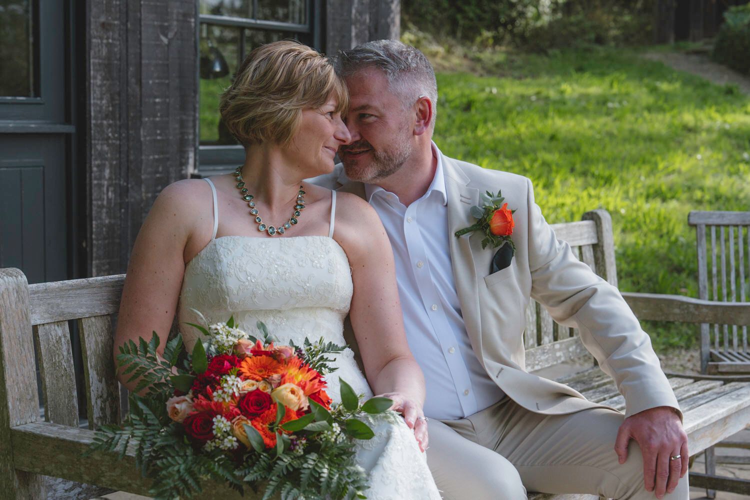 Bride and groom sitting on wooden bench, sharing a romantic moment, bride holding vibrant orange and red bouquet.
