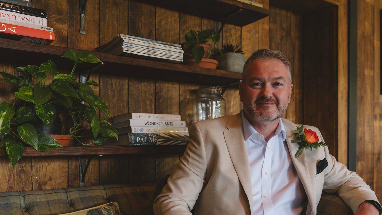 Smiling man in beige blazer with orange boutonniere seated near rustic wooden shelves with books and greenery.
