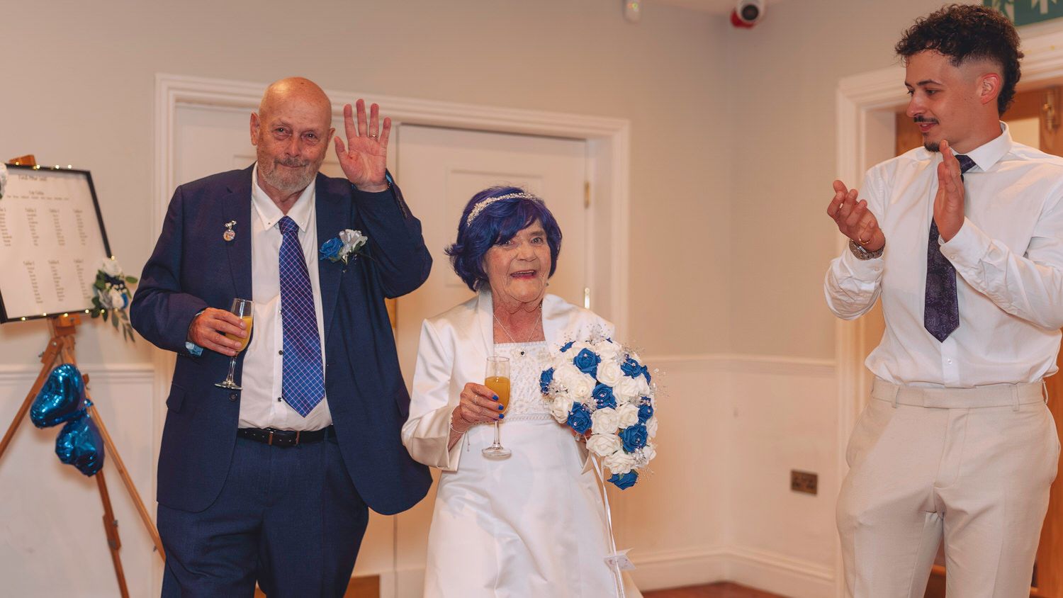 Elderly couple celebrating wedding anniversary in blue outfits, waving to guests in a decorated reception hall.