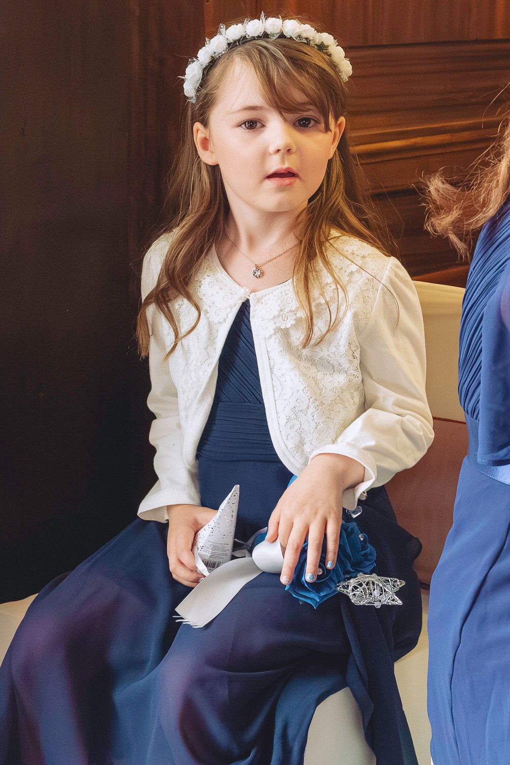 Young girl in navy dress, white lace jacket, and floral crown sits holding wedding accessories.