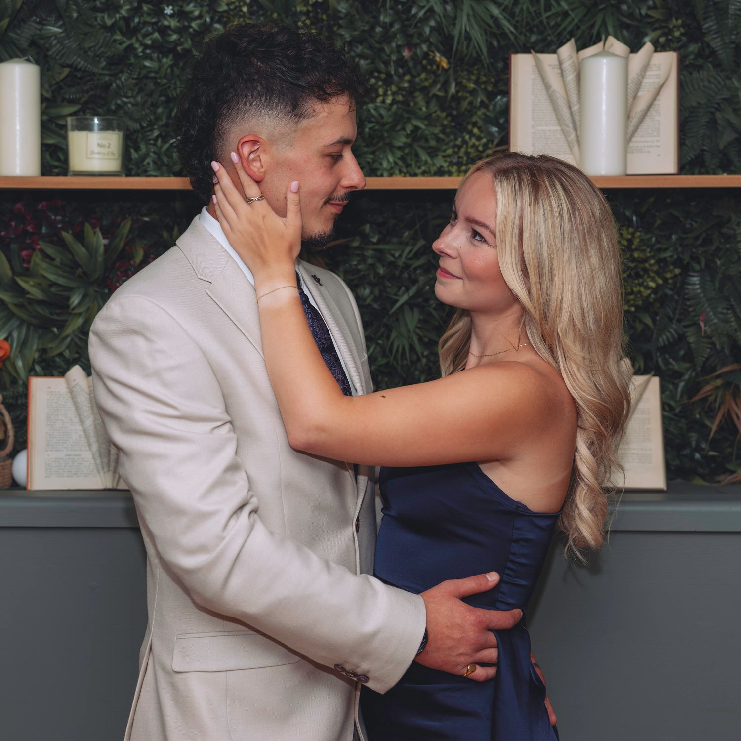 A couple in formal attire embraces tenderly in front of a lush green wall with decorative shelves.