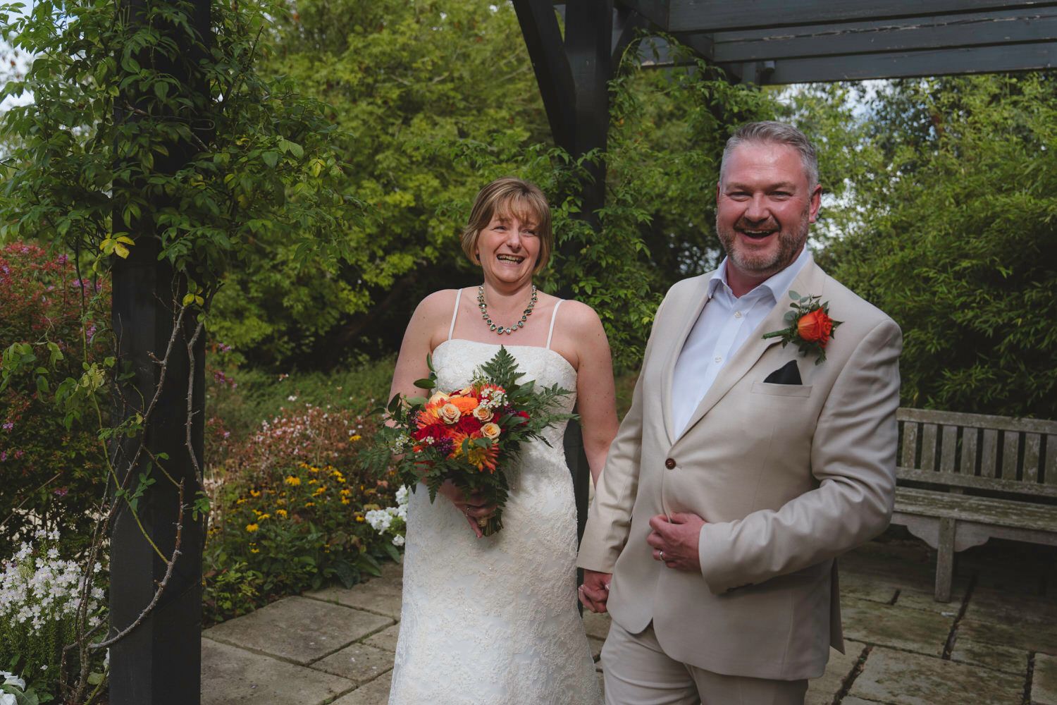 Happy bride and groom laughing together in a garden, bride holding colorful bouquet, groom in beige suit.