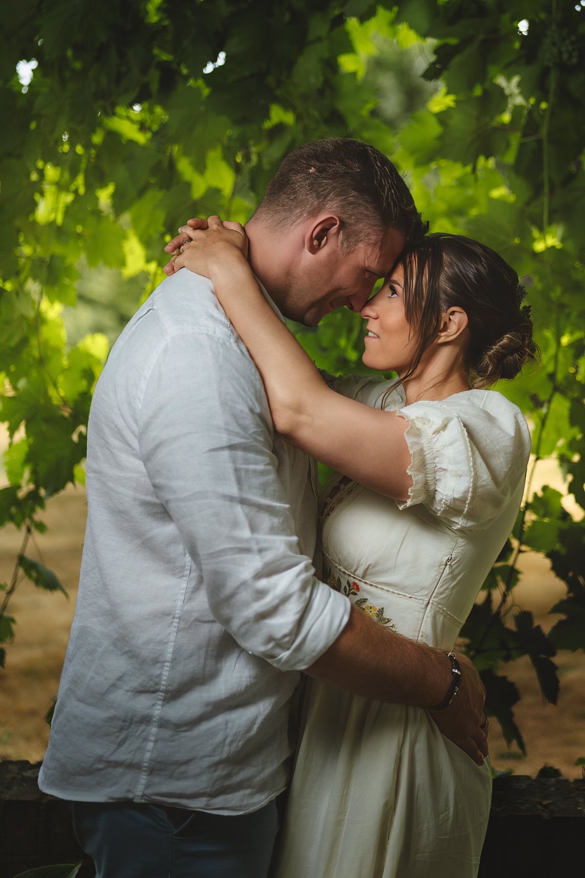 Couple embracing tenderly under lush green grapevine canopy in romantic outdoor setting.