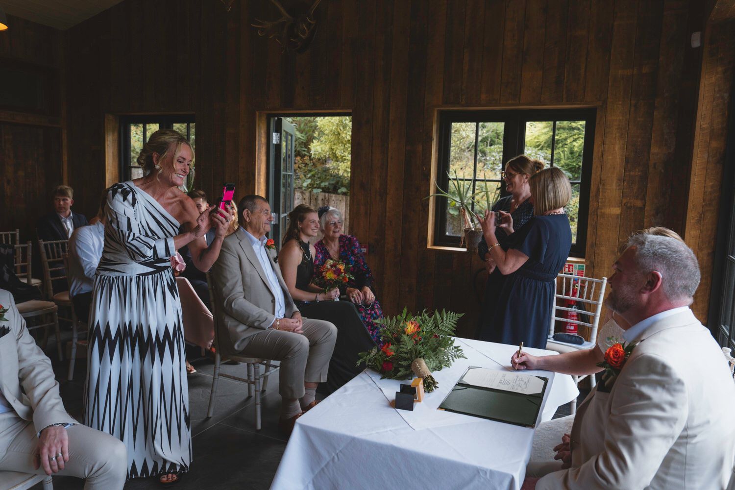 Wedding ceremony in rustic wooden venue with guests seated and standing, capturing moments on phones.