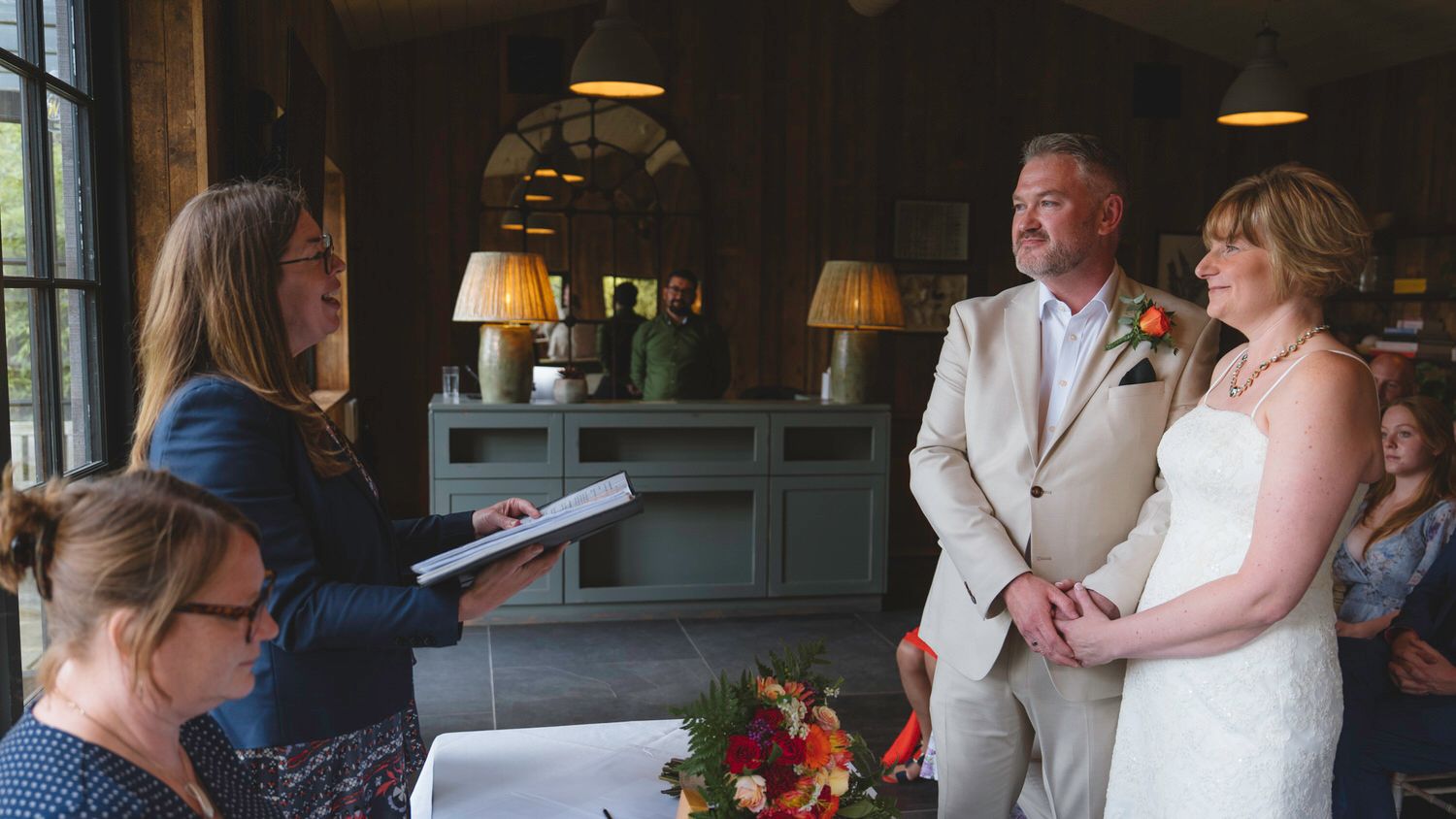 Couple exchanging wedding vows before an officiant in a rustic venue, bride in white gown, groom in beige suit.