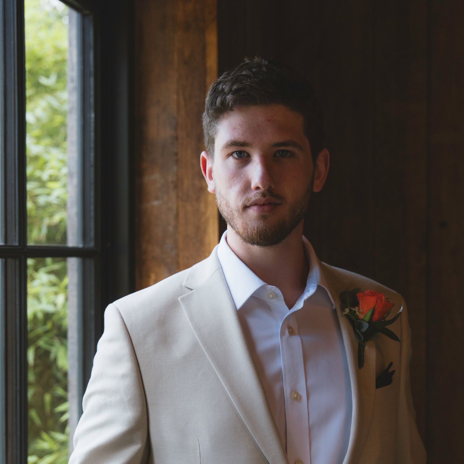 Young man in cream suit with boutonniere poses near window, likely a groom on his wedding day.