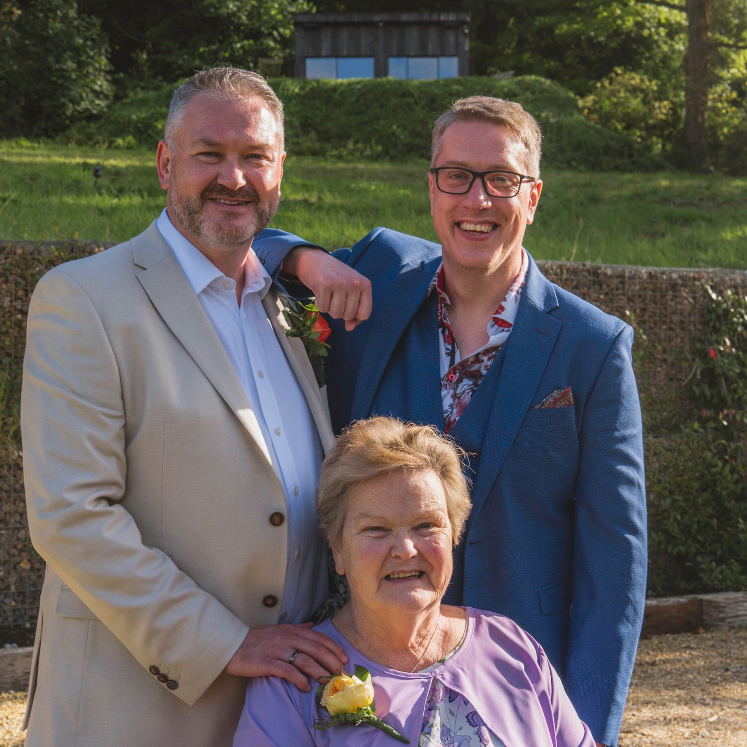 Three smiling people pose outdoors at a formal event, two men and an elderly woman in a purple dress.