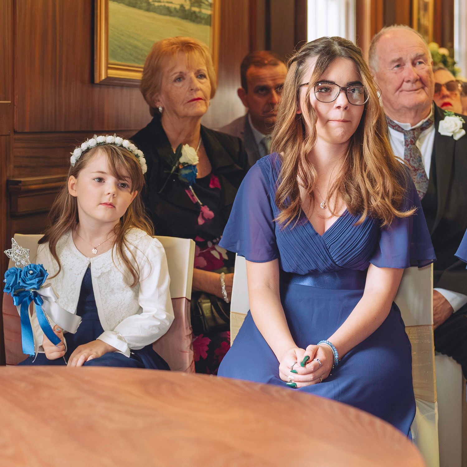 Women and girl in blue outfits seated at a wedding ceremony in an elegant room.
