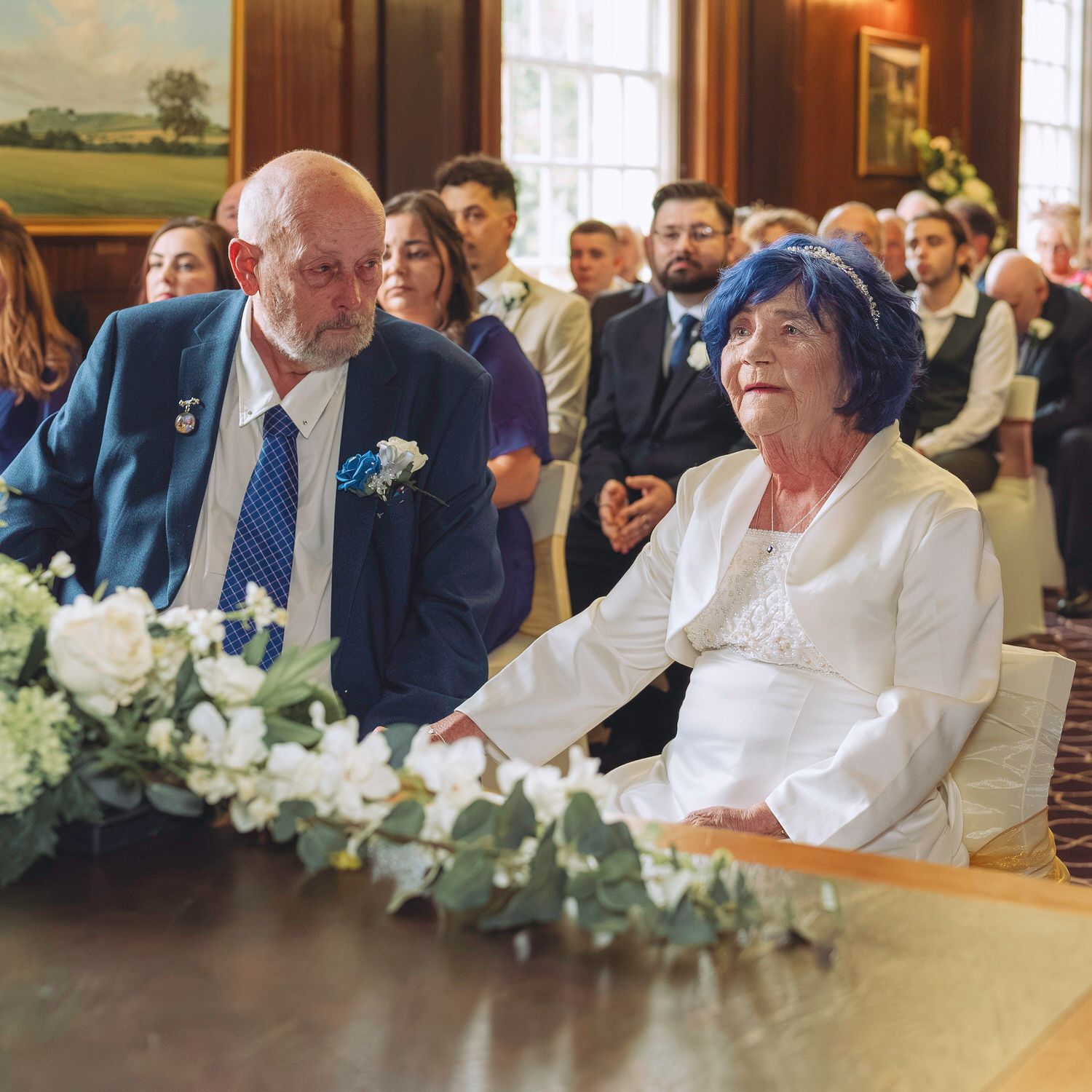 Elderly couple smiling at a wedding ceremony, man in blue suit with boutonniere, woman in white jacket with blue hair.