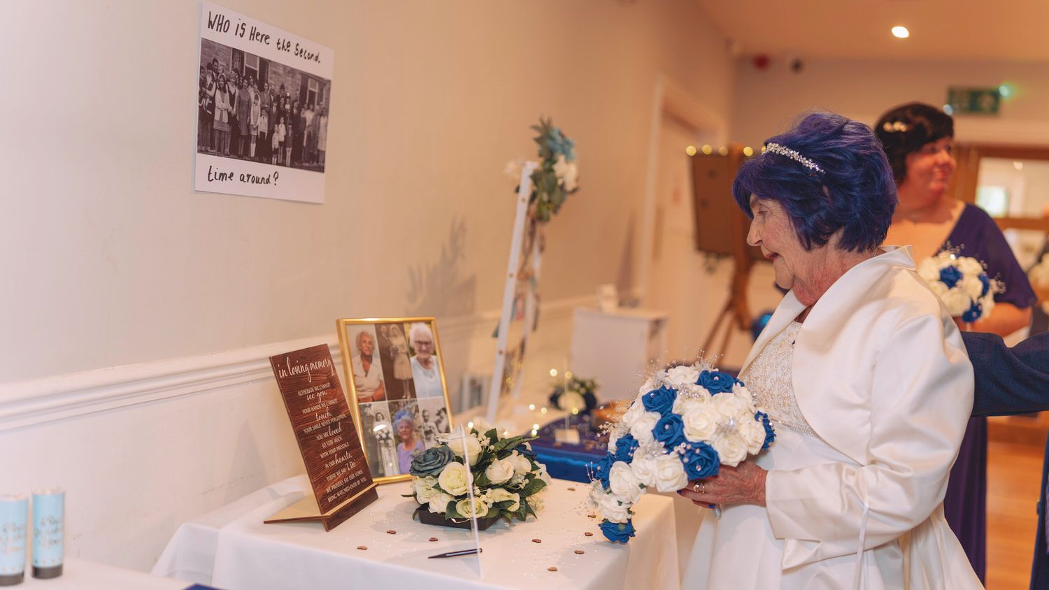 Elderly bride in white gown holds blue and white bouquet at wedding memory table with photos and floral decorations.