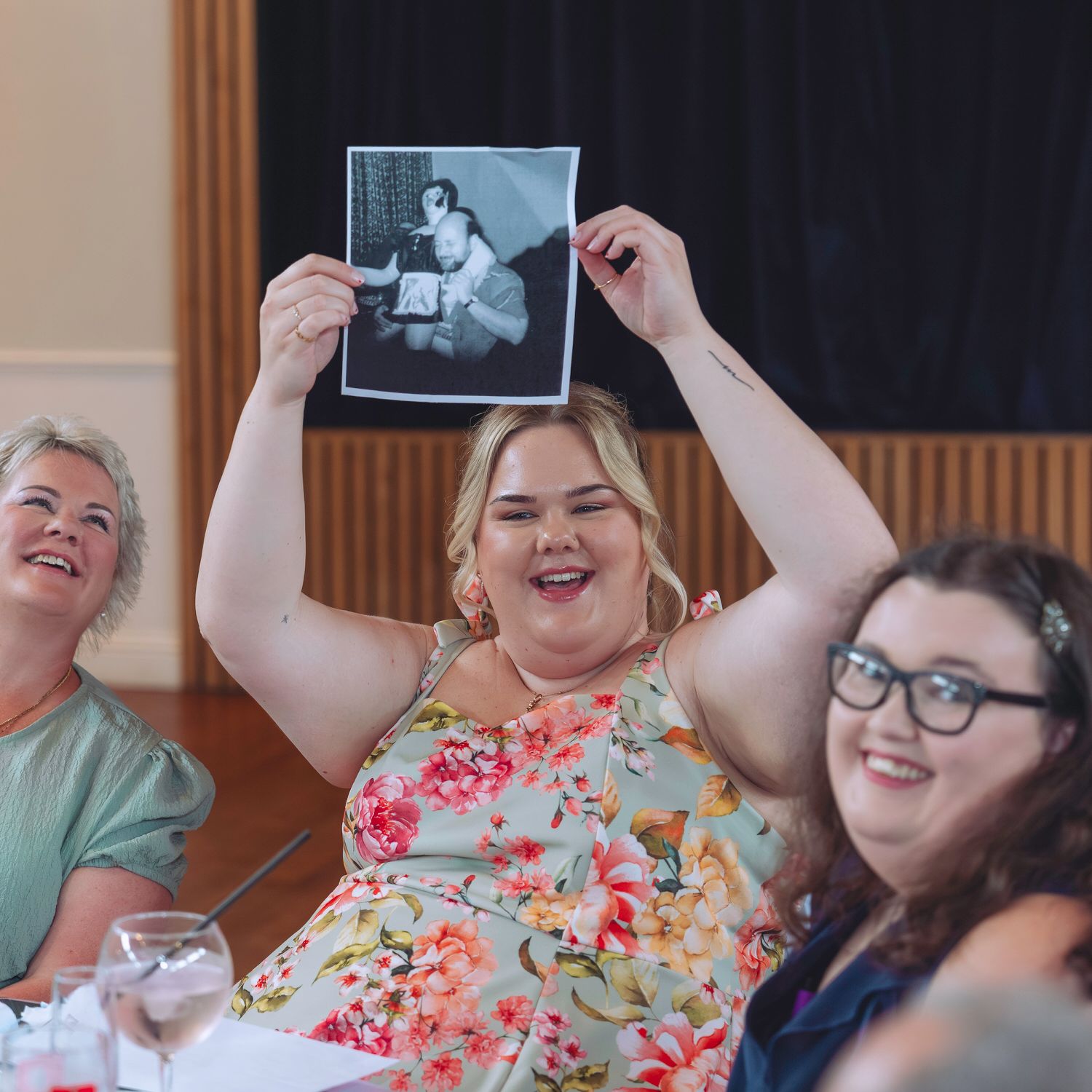 Woman in floral dress holds up black and white photo at a celebration, laughing with friends at a banquet table.