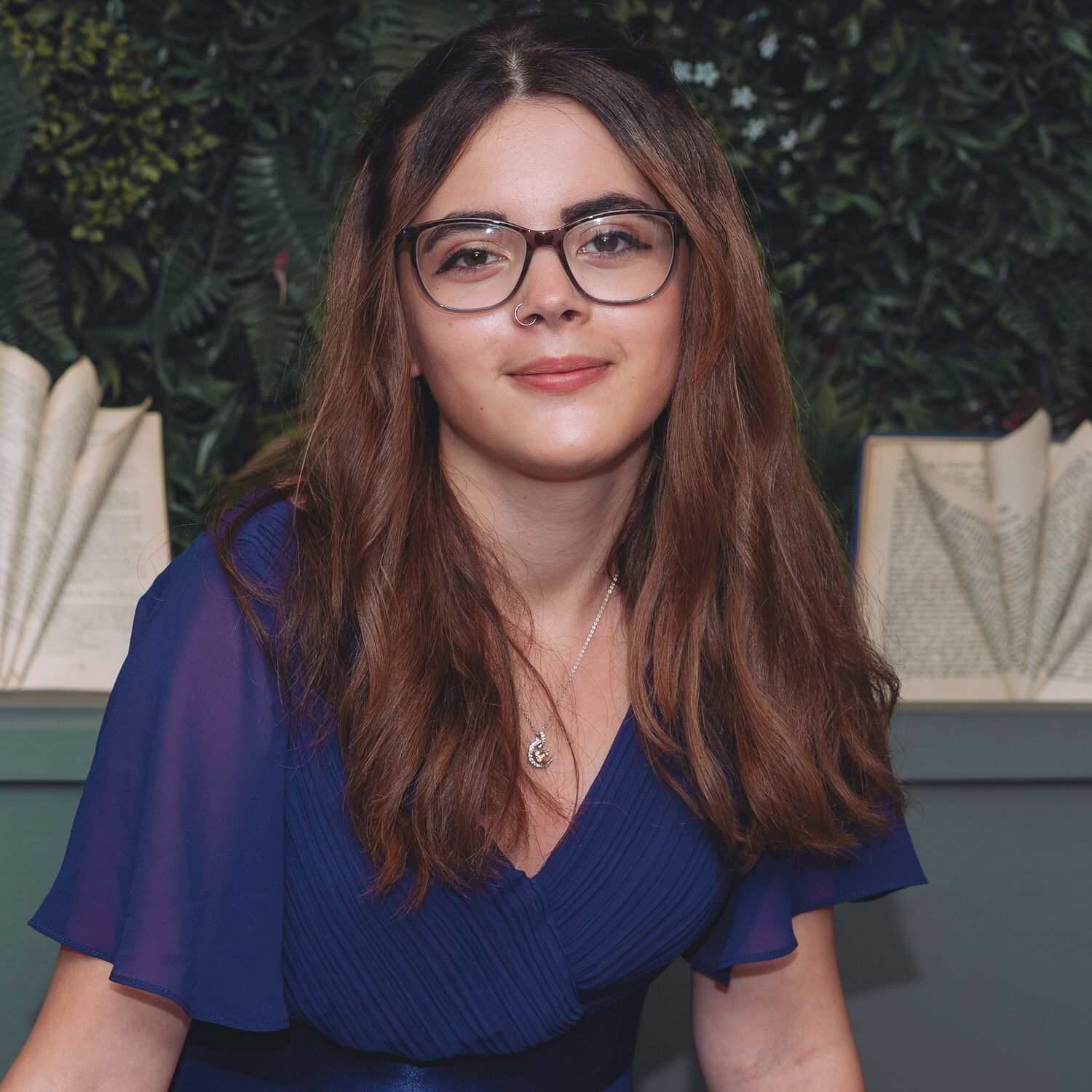 Young woman with glasses and long brown hair wearing a blue dress smiles in front of green foliage.