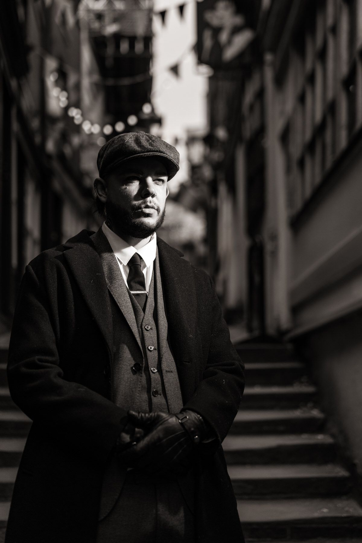 Man in vintage flat cap and long coat poses in moody black and white alleyway portrait.