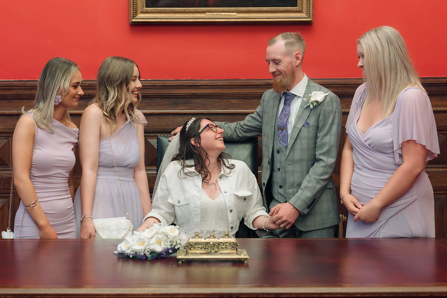 Bride and groom signing wedding register with three bridesmaids in lilac dresses in elegant red room.