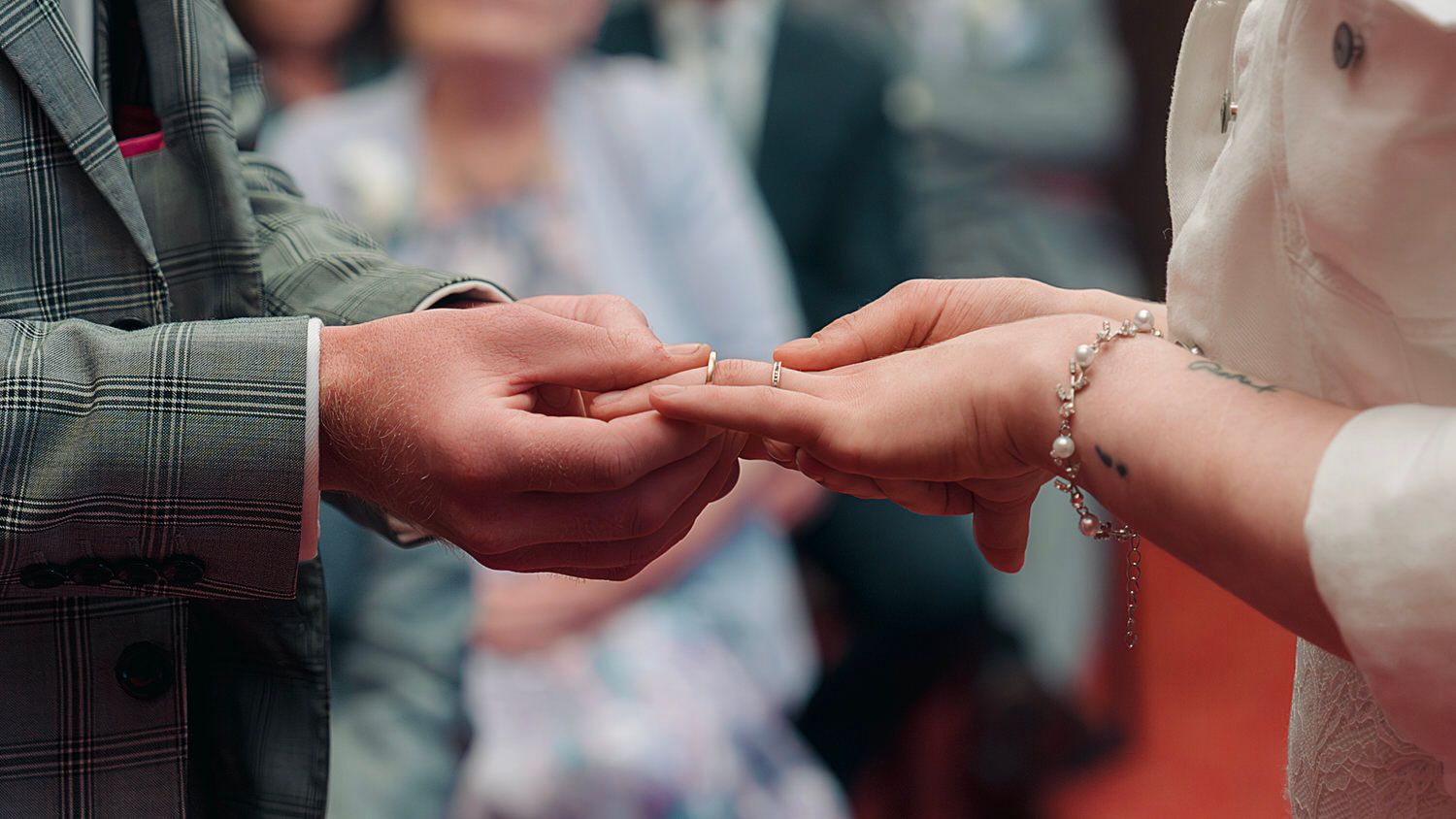 Groom placing a wedding ring on bride's finger during a ceremony, with guests visible in the background.