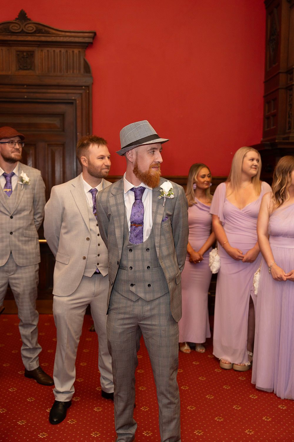 Groom in gray checked suit and fedora hat stands with groomsmen and bridesmaids in lavender dresses.