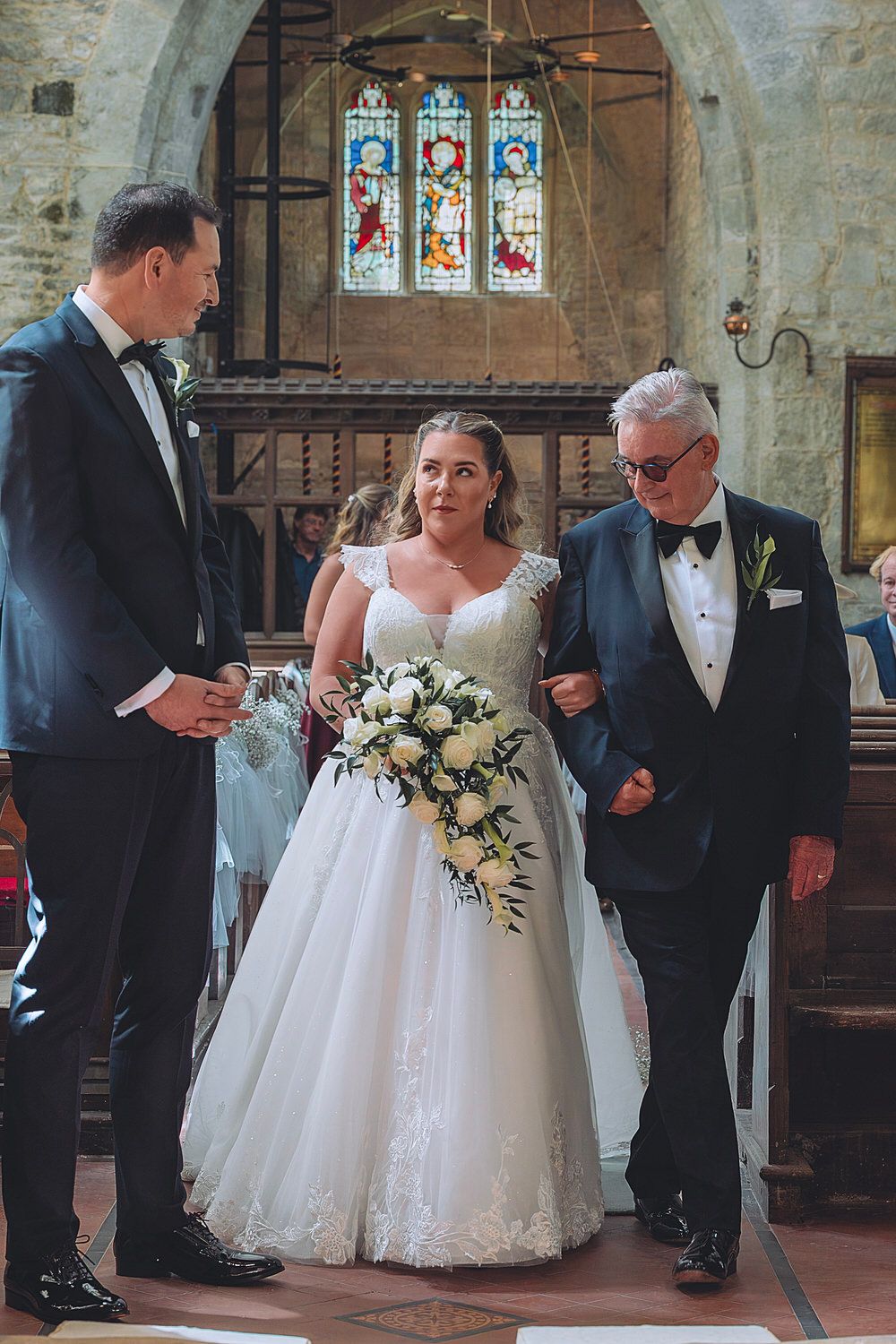 Bride in white lace ball gown walks down church aisle with escort, groom waiting, stained glass windows behind.