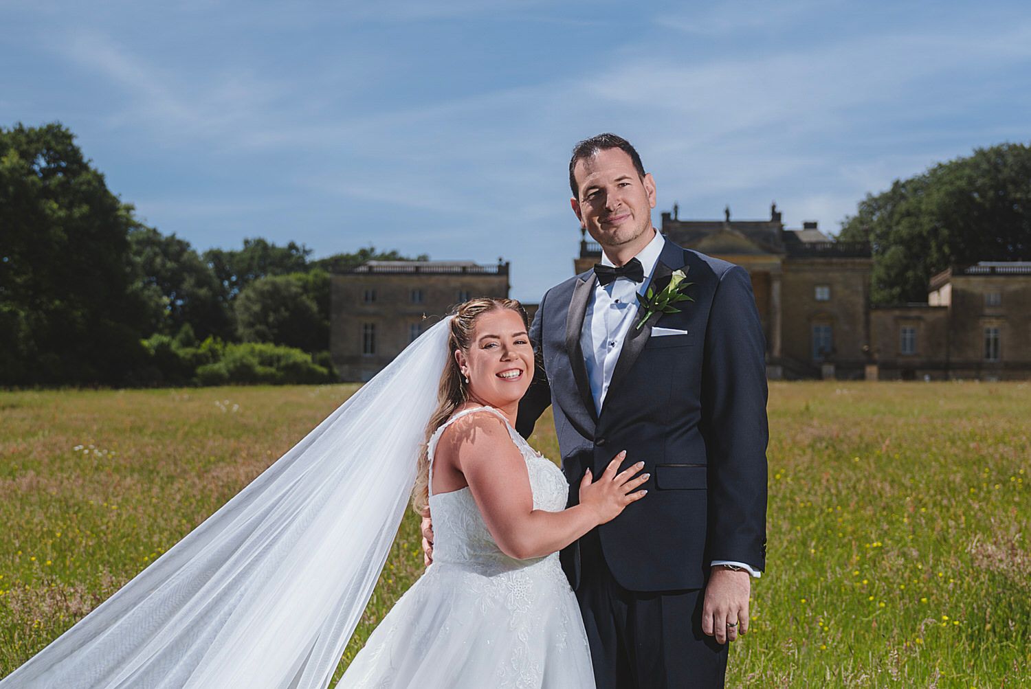 Bride and groom smiling together in a green meadow with a historic manor house in the background on their wedding day.