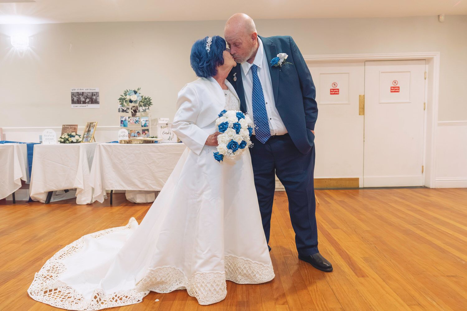 Bride in white gown with blue hair kisses groom in navy suit holding teal bouquet at wedding reception.