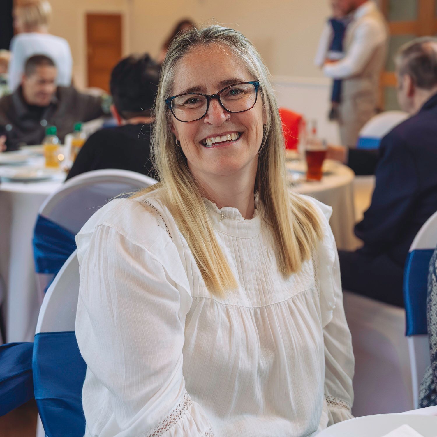 Smiling blonde woman with glasses in white blouse seated at a banquet event with guests dining in background.
