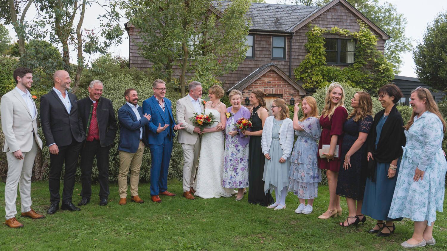 Wedding party posing outdoors on green lawn beside brick house, bride in white gown surrounded by guests in formal attire.