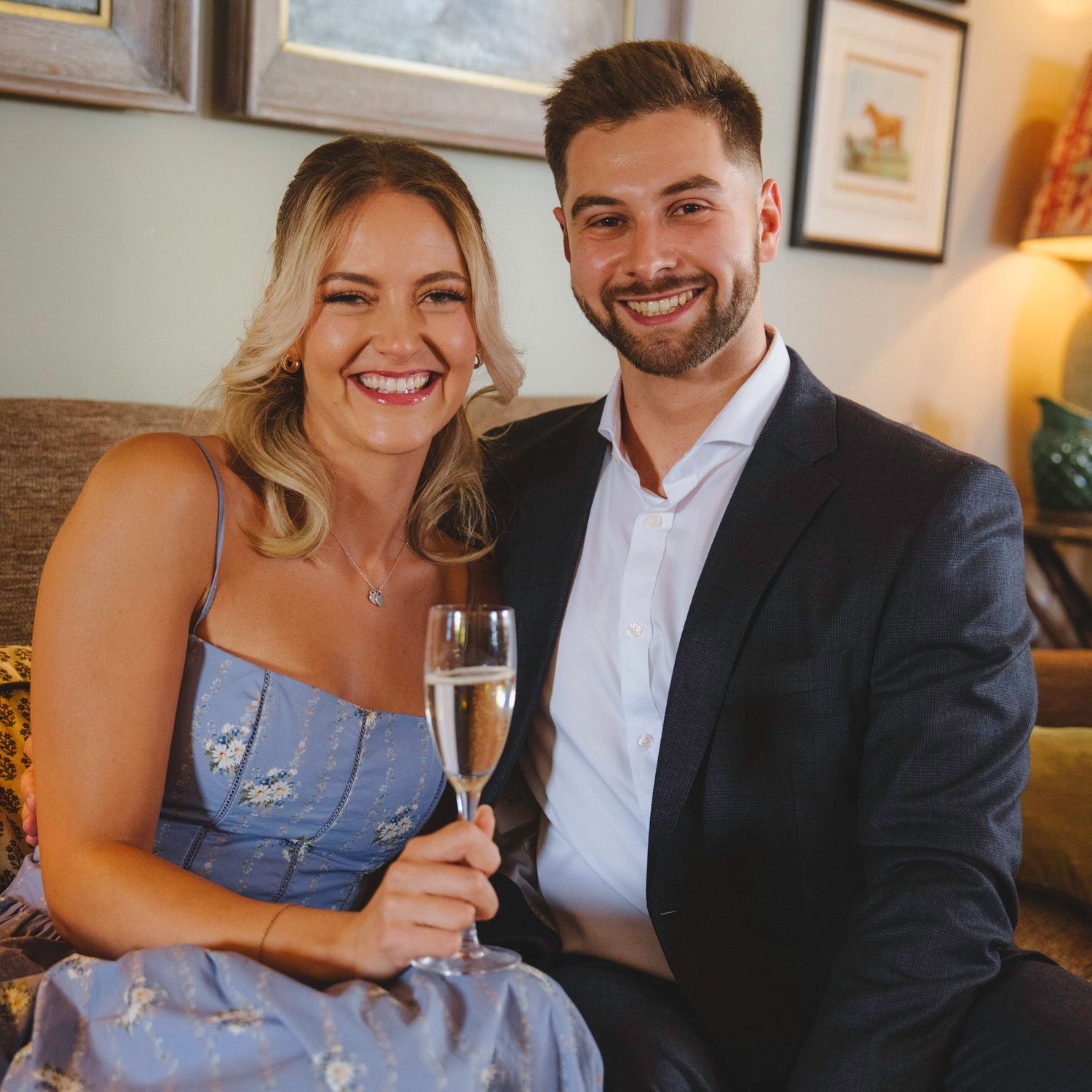 Smiling couple celebrating with champagne glasses, woman in blue dress and man in dark blazer sitting together.