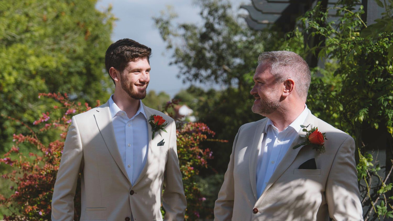 Two grooms in beige suits with red boutonnieres share a smile outdoors at a garden wedding ceremony.