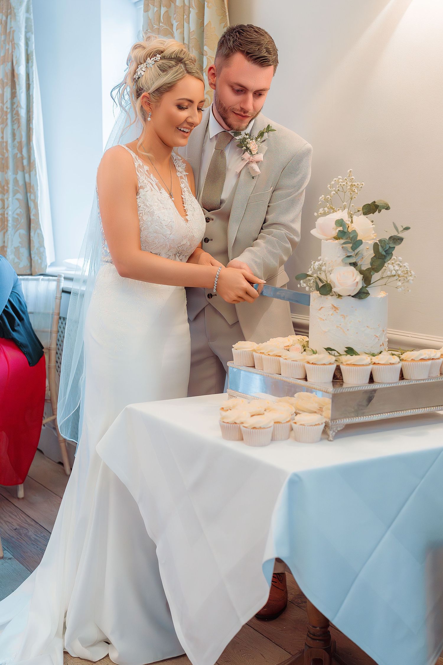 Bride and groom cutting their white wedding cake together, surrounded by cupcakes on a decorated table.