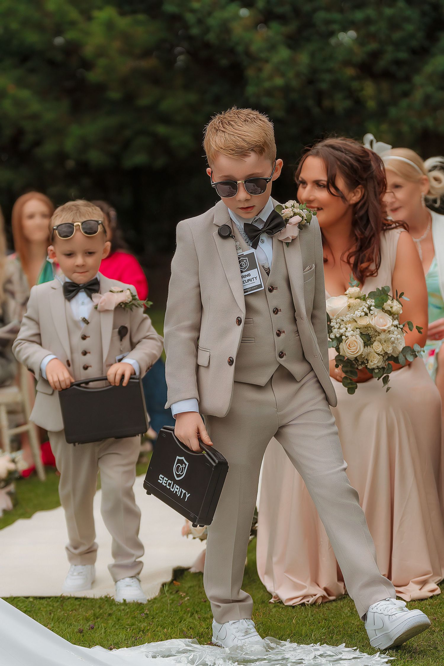 Two young ring bearers in tan suits and sunglasses carry security briefcases at an outdoor wedding ceremony.