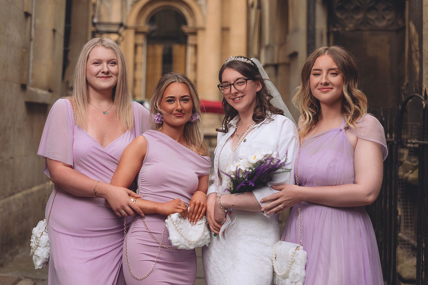 Bride in white gown holds bouquet with three bridesmaids in lavender dresses posing outdoors near stone building.