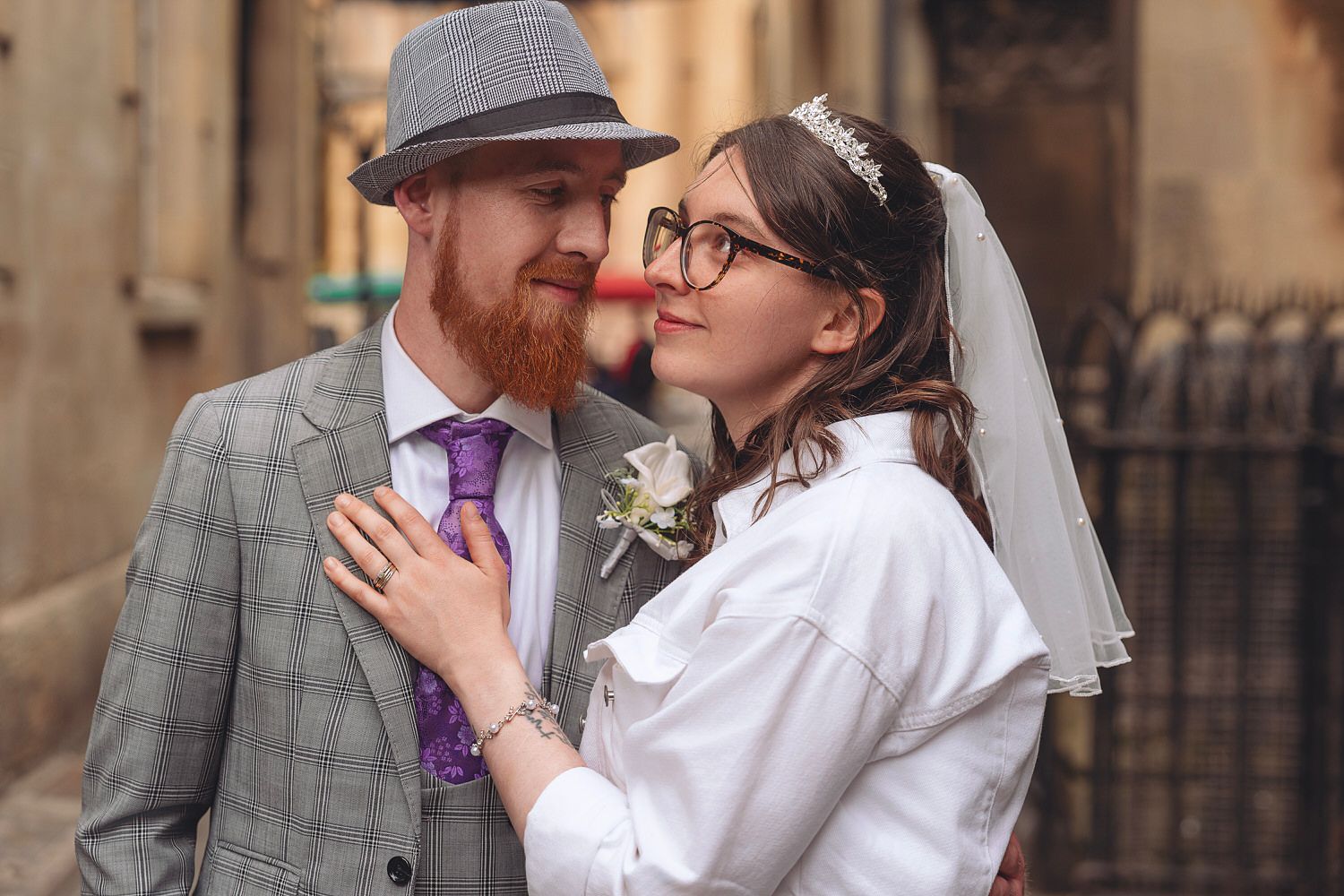 A bride and groom share a romantic moment, she in a white dress and veil, he in a grey suit with purple tie.